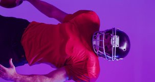 Vertical video of caucasian male american football player holding ball, with neon purple lighting. Sport, movement, training and active lifestyle concept.