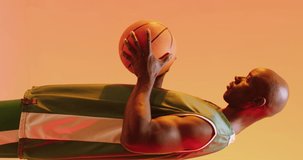 Vertical video of african american male basketball player holding ball on orange background. Sports and competition concept.