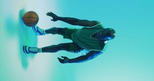 Vertical video of african american male basketball player bouncing ball on green background. Sports and competition concept.