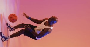 Vertical video of african american male basketball player throwing ball on pink background. Sports and competition concept.