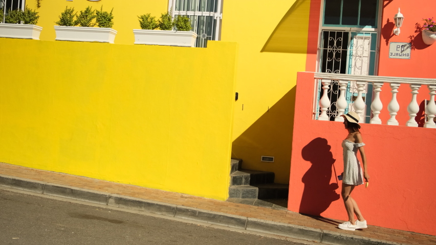 woman in White dress dancing on the street. Joyful african girl in summer dress turning around. stylish girl traveler in a dress holds the flag of south africa and has fun near the colored houses
