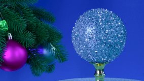 Near the large decorated rotating Christmas tree branches, a Christmas ball rotates on a blue background