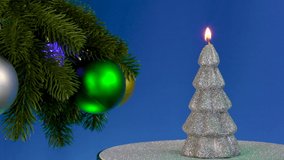 Near the large decorated rotating Christmas tree branches, a New Year's candle rotates on a blue background