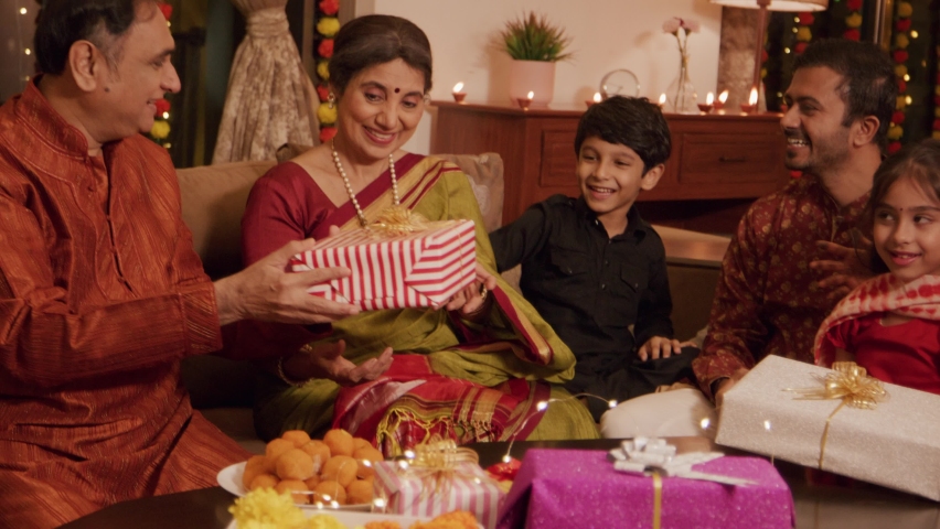Ethnic Hindu Indian happy grandparents are exchanging or giving a gift box to a young cute smiling grandson with a whole family sitting together and celebrating Diwali festival in a decorated home. Royalty-Free Stock Footage #1095035465