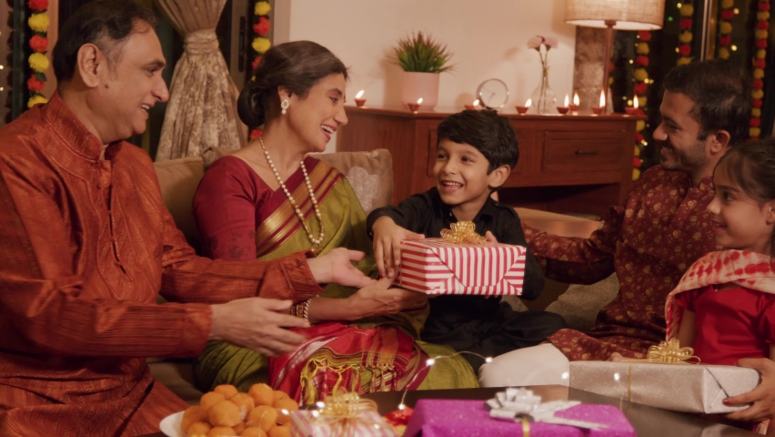 Ethnic Hindu Indian happy grandparents are exchanging or giving a gift box to a young cute smiling grandson with a whole family sitting together and celebrating Diwali festival in a decorated home. Royalty-Free Stock Footage #1095035465