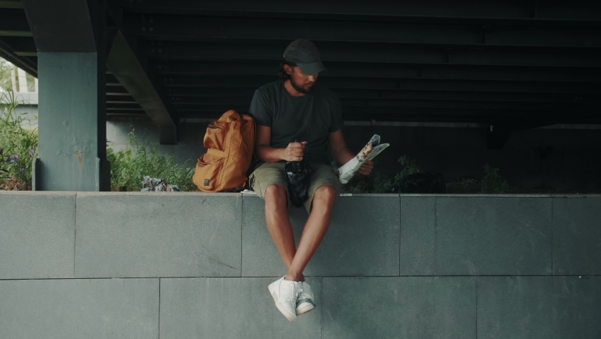 Tired homeless man, refugee tourist on a bench in the park drinking beer or wine, with backpack. Crisis, immigration and unemployment.