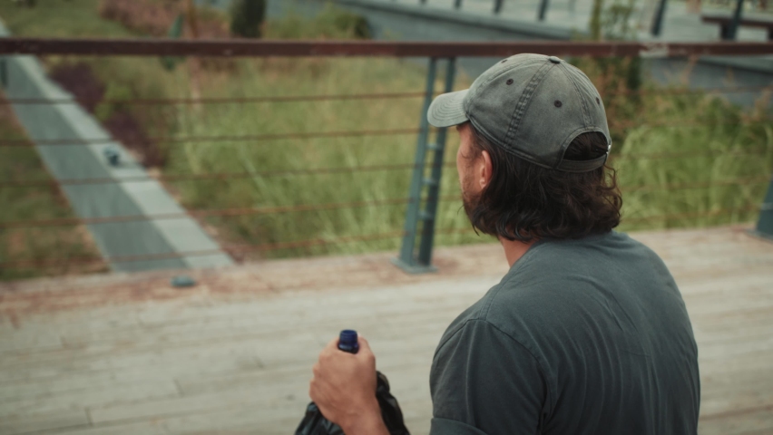 Tired homeless man, refugee tourist on a bench in the park drinking beer or wine, with backpack. Crisis, immigration and unemployment.