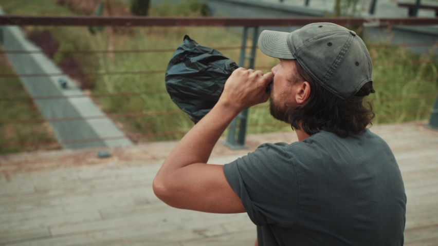 Tired homeless man, refugee tourist on a bench in the park drinking beer or wine, with backpack. Crisis, immigration and unemployment.