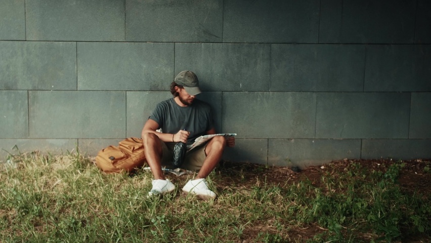 Tired homeless man, refugee tourist on a bench in the park drinking beer or wine, with backpack. Crisis, immigration and unemployment.