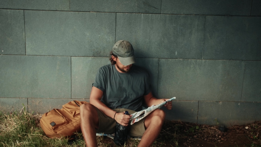 Tired homeless man, refugee tourist on a bench in the park drinking beer or wine, with backpack. Crisis, immigration and unemployment.
