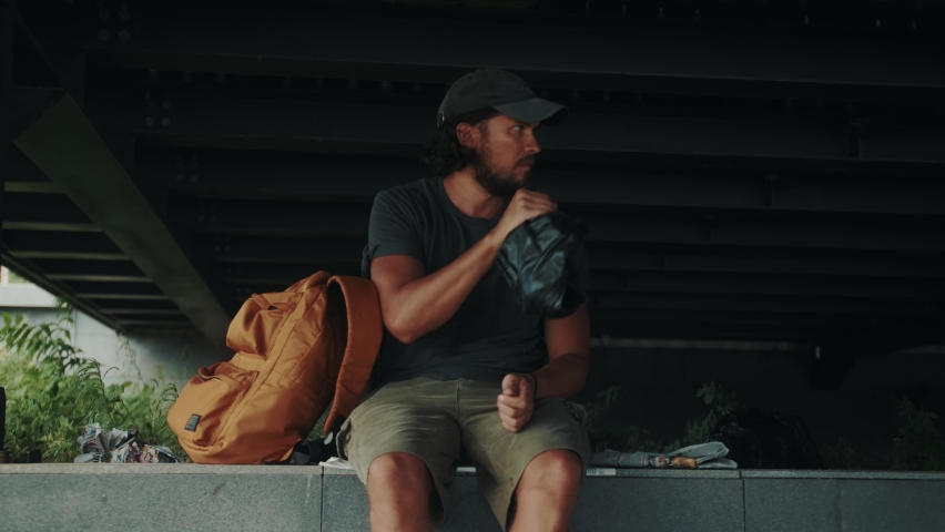 Tired homeless man, refugee tourist on a bench in the park drinking beer or wine, with backpack. Crisis, immigration and unemployment.