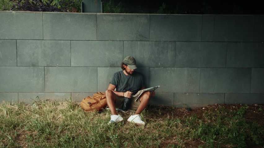 Tired homeless man, refugee tourist on a bench in the park drinking beer or wine, with backpack. Crisis, immigration and unemployment.