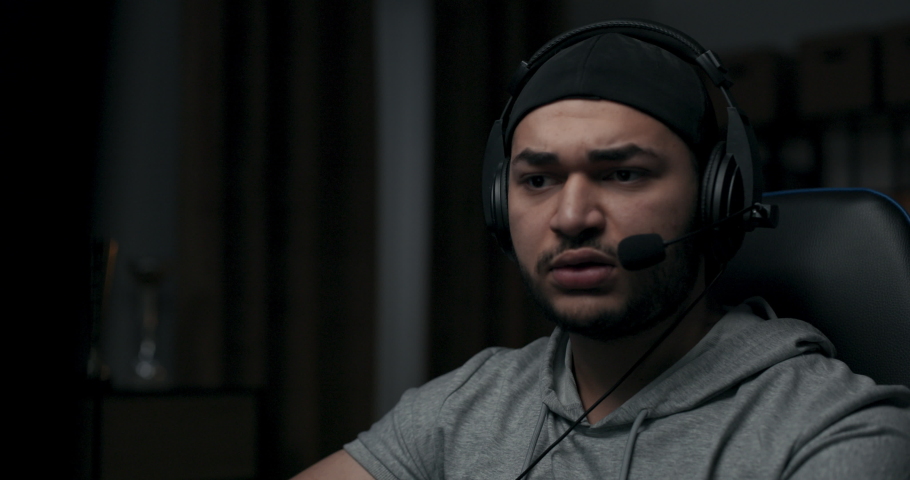 Close-up of bearded man concentrating on playing online game on computer. Guy is wearing cap and headphones. He is wearing gray T-shirt. Player sits in room, communicates with opponents in game.