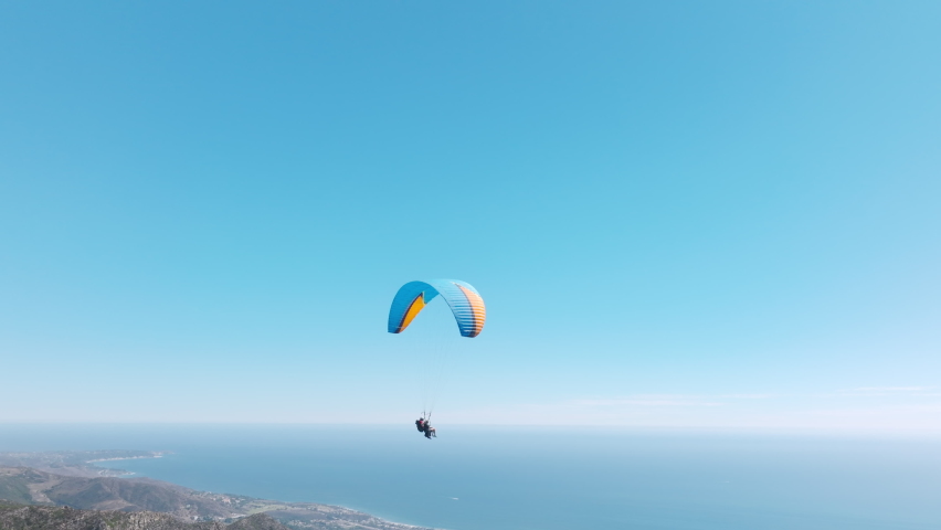 Slow motion up in the air, cinematic aerial drone footage above Malibu USA. Extreme paraglider flying against a clear blue sky, sunbeam shines into camera. Man with instructor flies with paraglide 4k Royalty-Free Stock Footage #1095043051