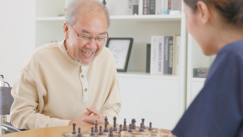 At home, a senior retiree plays chess with an Asian beauty nurse. Senior Asian man and his intelligent and thoughtful assistant girl playing chess together. Retirement man's leisure pastime On weekend Royalty-Free Stock Footage #1095048767