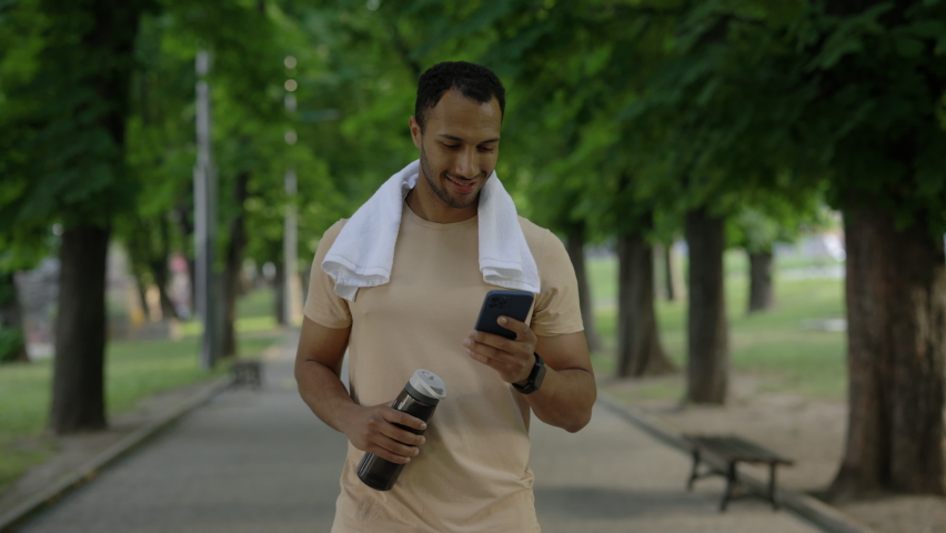Male african american runner is walking in the park after running using smartphone holding sport bottle and carries a towel around his neck wearing a smart watch on his arm Royalty-Free Stock Footage #1095050987