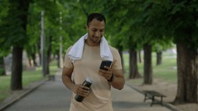 Male african american runner is walking in the park after running using smartphone holding sport bottle and carries a towel around his neck wearing a smart watch on his arm - Powered by Shutterstock - Get 15% off with code: PIKWIZARD15