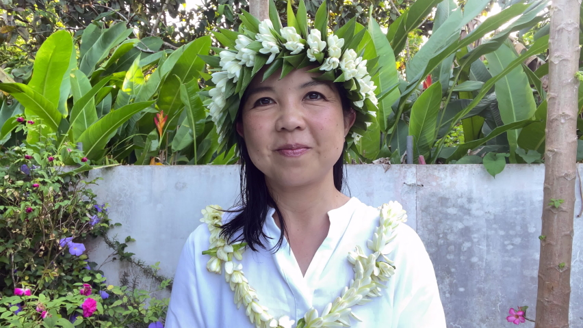 An asian woman wearing a flower crown and necklace in a tropical garden laughing 