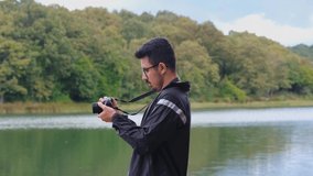 Young man filming lake with professional camera, taking video concept, 4k 60 fps, glasses man using camera in nature, beautiful landscape with cloudy sky, activity and leisure in forest, one person