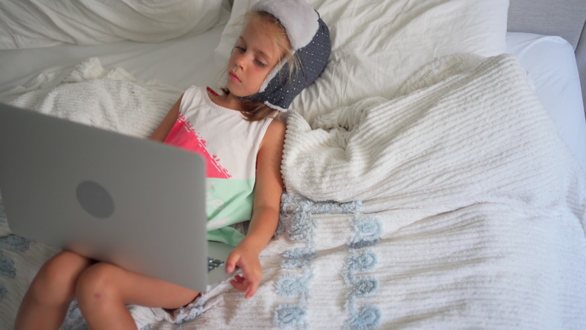 Child in funny hat watching video on laptop lies sofa at home. Little caucasian girl enjoy cartoons on laptop sitting bed in bedroom. Joyful preschool kid girl use laptop computer