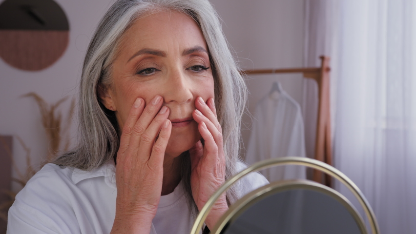 Senior gray-haired older 60s woman 50s lady granny with wrinkled beautiful aging face with perfect smooth skin looking at mirror reflection touching cheeks massaging skincare cosmetology treatment Royalty-Free Stock Footage #1095073037