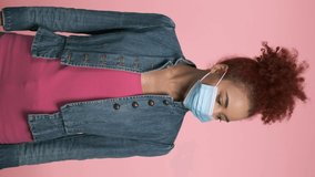 Vertical video of young african american woman posing in medical protective mask on pink studio background. Concept of health care, virus protection, pandemic