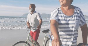 Animation of merry christmas text over happy diverse senior couple with bikes on sunny beach. Christmas, holidays, festivity, tradition and celebration concept digitally generated video.