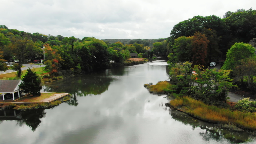 Aerial view of lake surrounded with houses and green forest of threes during summer cloudy day. Flying by drone over the river. High quality 4k footage