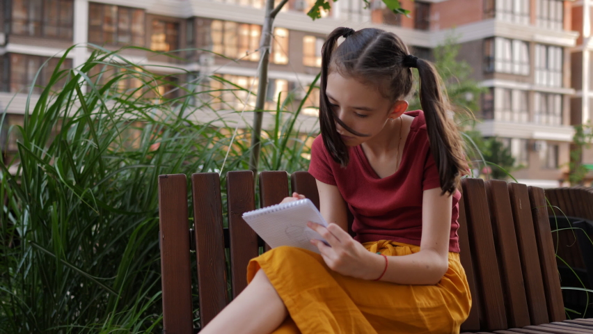 A creative child girl draws in the yard of the house in her notebook.