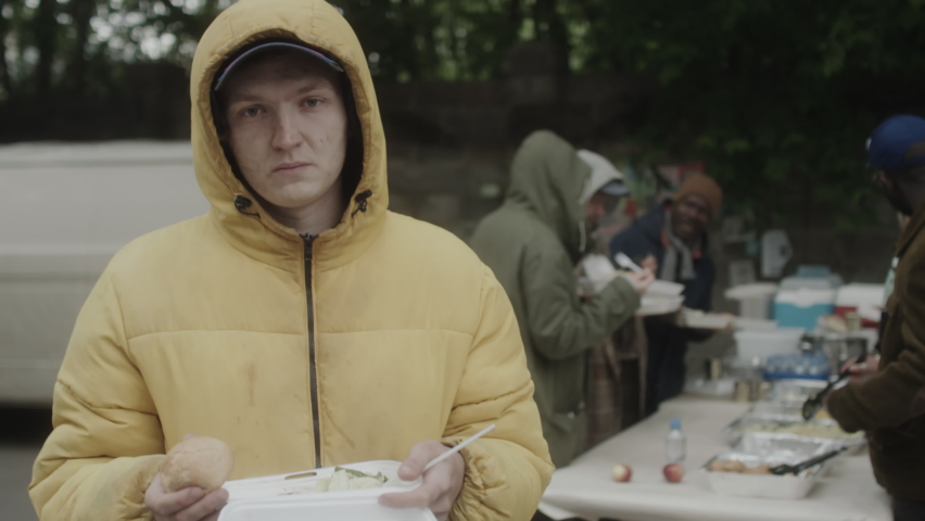 Portrait of young homeless man in outerwear holding free food in container and posing for camera near outside soup kitchen
