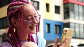 Portrait of a smiling caucasian teenage hipster girl with pink braids using a smartphone on a street background.Summer concept.Generation Z style.Social media concept.