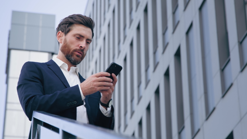 Stylish handsome businessman holding phone reading email stands on street smiling writes message communicates in social network pensive man browses goods in online store using mobile app on smartphone | Shutterstock HD Video #1095108479