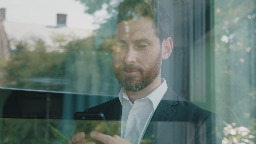 Close up reflection in glassy window blue glass Caucasian bearded 40s middle-aged man adult businessman walking indoor in office company browsing scrolling phone mobile app looking outside pensive - Powered by Shutterstock - Get 15% off with code: PIKWIZARD15
