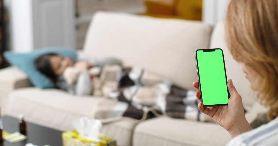 Caucasian female holding smartphone with green screen in hand while sitting at home. Woman in room holds cellphone with chroma key and medicines. Sick little girl laying on sofa indoor. Health concept