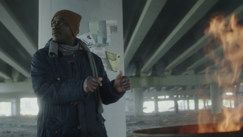 African American homeless man talking with his friend while standing in abandoned building and heating their hands around fire barrel