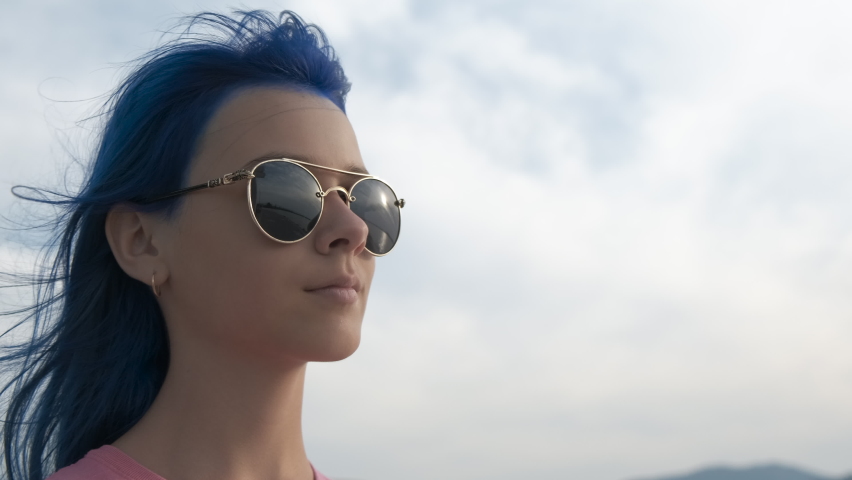 Teen with blue hair and glasses outdoor. A pretty young girl in sunglasses with dyed hair relax in the sea wind on shore. A concept of summer relaxation during sea vacation.