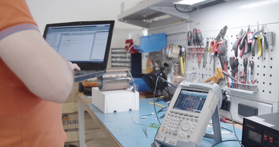 Electronics engineer programming software while holding the laptop in his arm next to a spectrum analyzer in electronics laboratory | Shutterstock HD Video #1095117975