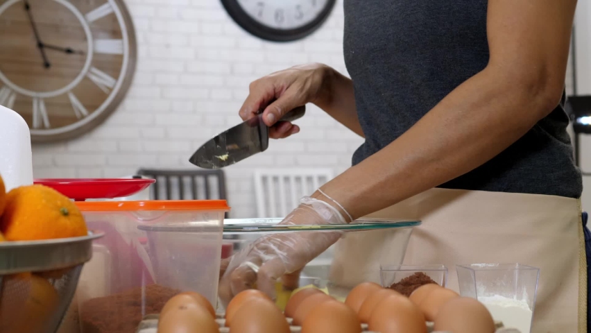 Middle aged woman 50 year old make homemade bakery. Adult woman wearing glove hand cutting butter. People Lifestyle old female cooking in kitchen. 