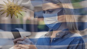 Animation of flag of uruguay waving over caucasian woman wearing face mask in city street. global health precautions during covid 19 pandemic concept digitally generated video.