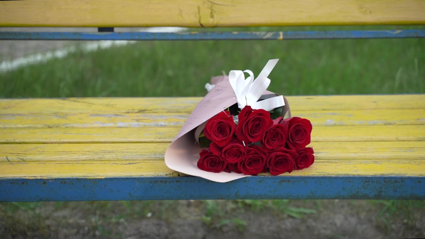 Thrown decorated bouquet of nine red roses lies on yellow-blue old shabby bench on green grass. Wind blows, leaves and petals on the cut flowers sway. | Shutterstock HD Video #1095126787