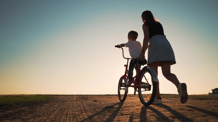 Happy family concept. Mom teaches her son to ride bike on green grass. Child rides bicycle along rural road. Green energy. Mom teaches her son to ride bike for the first time in park at sunset. Royalty-Free Stock Footage #1095127553