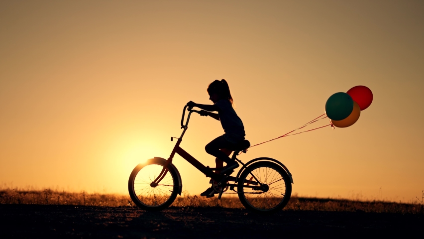 Dream kid. Silhouette of kid on bike in park. Girl rides in park on green grass. Child games in nature.Traveling with balloons on bike.Active child freedom in summer.Girl learns to ride bike in nature Royalty-Free Stock Footage #1095127555