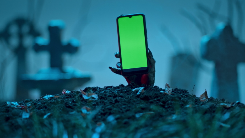 Zombie hand holds smartphone with green screen out of grave. Holiday event Halloween concept. Royalty-Free Stock Footage #1095127619