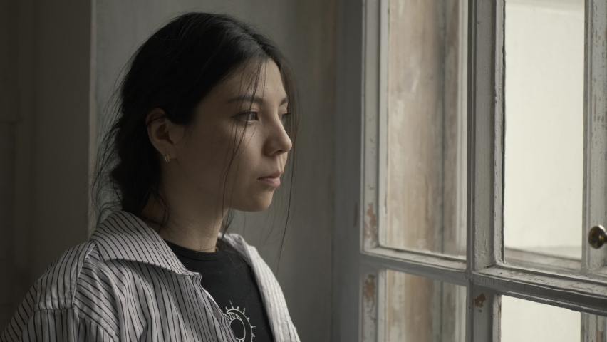 Asian Sad young woman standing by the window and sitting on a chair, looks unhappy, lost due to unpleasant thoughts, suffering from life problems. Filmed in cinematic style
