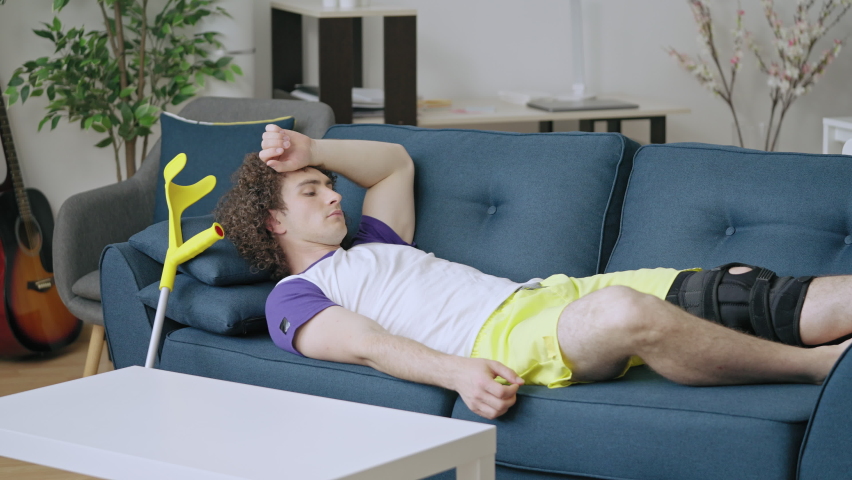 Young man lying on couch, rubbing injured knee in immobilizer, suffering pain