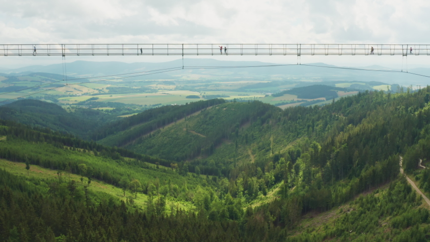 Suspension Sky Bridge with tourists high over forestry valley in highland in Dolni Morava. Mountain range on horizon under white cloudy sky aerial view