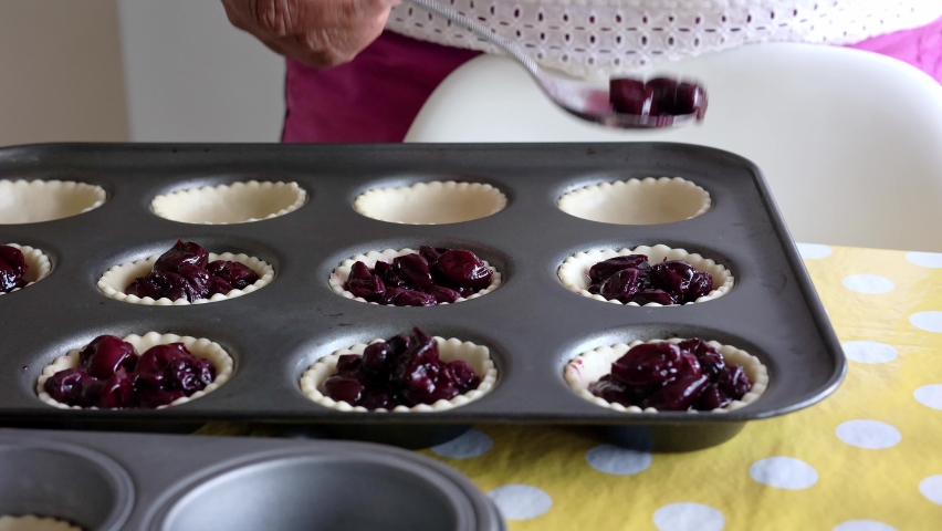 Woman making cherry tarts, filling pastry cases with stewed cherry compote, while an excited child runs into the kitchen.  Real time. Royalty-Free Stock Footage #1095146987