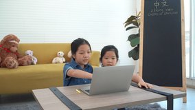 Two asian siblings learning chinese language online together with laptop on desk at home. Happy asian young girl knowledge language on notebook pc sitting together with yellow sofa,black board in room