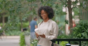 Attractive curly haired woman standing on the street with coffee and chatting in messenger, wearing glasses and light shirt