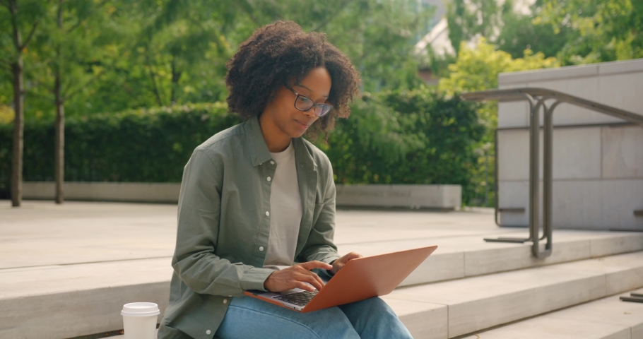Curly haired African American female student, seating on coffee break outdoors on the stairs and typing on her laptop computer with a joy smile